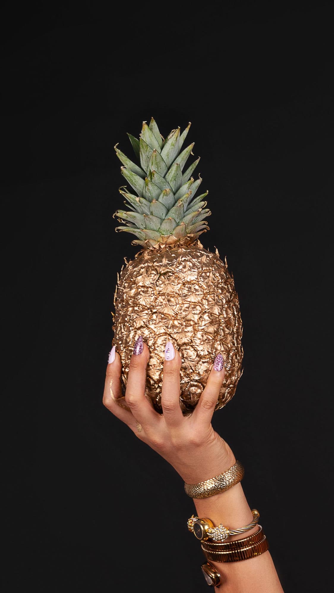 A manicured, gold-braceleted hand holding a gilded pineapple against a black backdrop