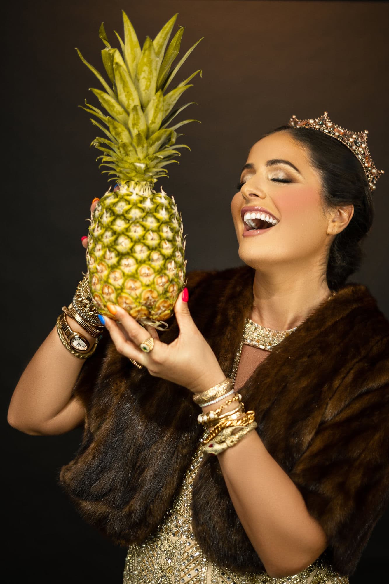 Charlotte laughing, wearing a crown and fur, holding up a pineapple in a warm editorial studio portrait